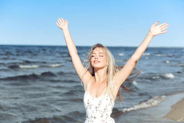 Szczęśliwa blondynka piękna kobieta na plaży oceanu stojąca w białej letniej sukience, podnosząc ręce.