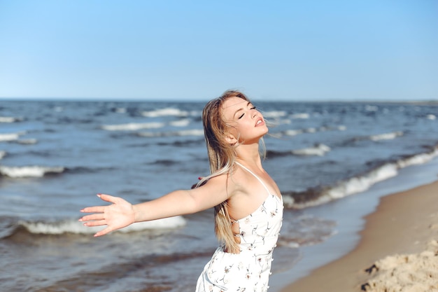 Szczęśliwa blondynka, piękna kobieta na plaży oceanu, stojąca w białej letniej sukience, podnosząc ręce.