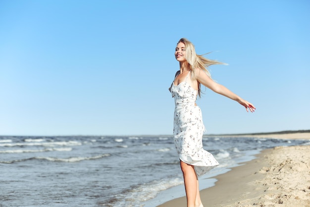 Szczęśliwa blondynka, piękna kobieta na plaży oceanu, stojąca w białej letniej sukience, z otwartymi ramionami.