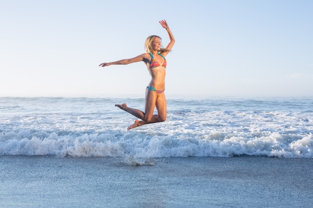 Szczęśliwa blondynka przeskakuje na plaży w bikini