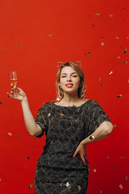 Szczęśliwa blondynka w błyszczących modnych ubraniach jest szczęśliwa trzymając kieliszek szampana i pozuje z konfetti na czerwonym tle