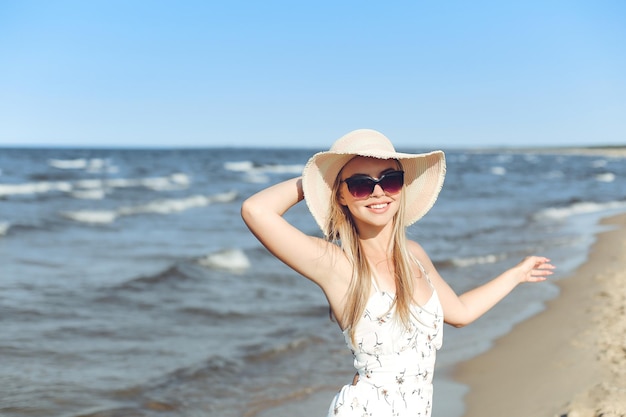 Szczęśliwa blondynka w wolnym szczęściu na plaży oceanu, stojąca w okularach przeciwsłonecznych i kapeluszu.