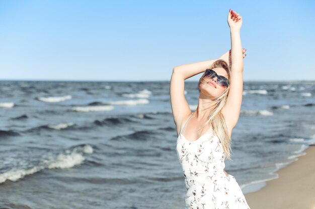 Szczęśliwa blondynka w wolnym szczęściu na plaży oceanu, stojąca w okularach przeciwsłonecznych.