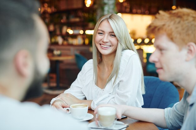 Szczęśliwa blondynka z uśmiechem toothy, mając filiżankę cappuccino podczas rozmowy z dwoma facetami przy stole w kawiarni po zajęciach