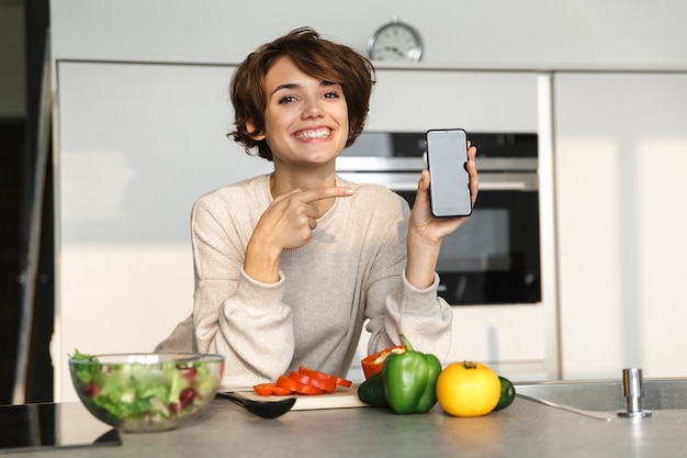 Szczęśliwa brunetka kobieta pokazuje pusty ekran smartfona i wskazuje na to podczas gotowania przy stole w kuchni