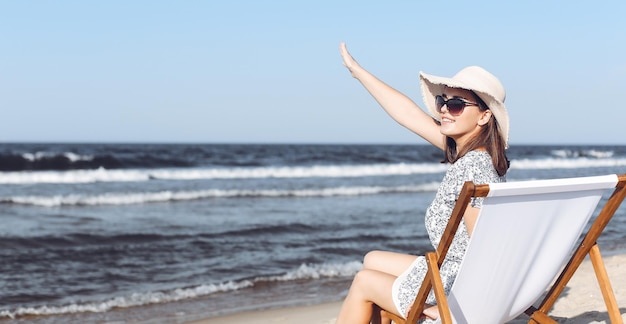 Szczęśliwa brunetka kobieta siedzi na drewnianym leżaku na plaży oceanu, machając i witając kogoś ręką.