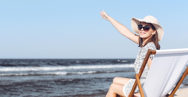 Szczęśliwa brunetka kobieta siedzi na drewnianym leżaku na plaży oceanu, machając i witając kogoś ręką