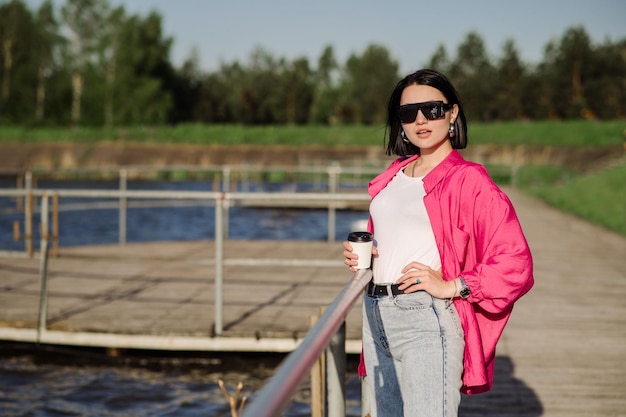 Szczęśliwa brunetka kobieta w okularach przeciwsłonecznych spacerująca w pobliżu jeziora na drewnianym molo w słoneczny dzień