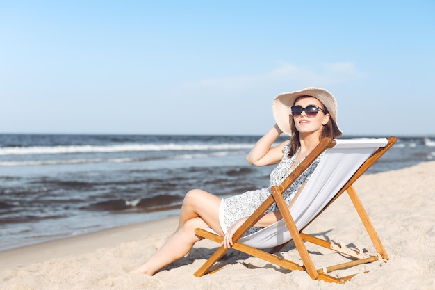 Szczęśliwa brunetka w okularach przeciwsłonecznych i kapeluszu relaksująca się na drewnianym leżaku na plaży oceanu.