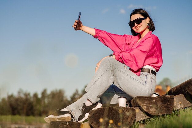 Szczęśliwa brunetka w okularach przeciwsłonecznych robi selfie za pomocą telefonu komórkowego nad błękitnym niebem na świeżym powietrzu w parku