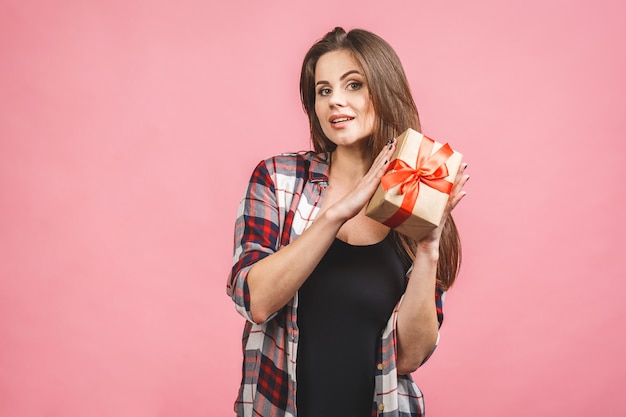 Szczęśliwa brunetki kobieta w przypadkowym mienie prezenta pudełku i patrzeć kamerę podczas gdy cieszy się nad różowym tłem