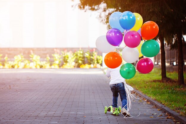 Szczęśliwa Chłopiec Bawić Się Z Wiązką Balony Outside I Jedzie Hulajnoga.