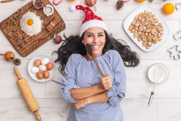 Szczęśliwa ciemnowłosa kobieta w świątecznym kapeluszu trzymająca drewnianą łyżkę i leżąca na ziemi z tradycyjnymi składnikami, takimi jak mąka, jajka, pomarańcze, a także formy do pieczenia, wałki lub pierniki.