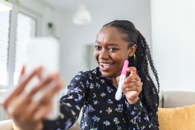 Szczęśliwa, Ciężarna Afrykanka Pokazuje Swój Test Ciążowy I Robi Selfie Podczas Wideorozmowy. Szczęśliwa Kobieta Biorąc Zdjęcie Testu Ciążowego Z Telefonu Komórkowego I Publikowanie Obrazu W Mediach Społecznościowych.