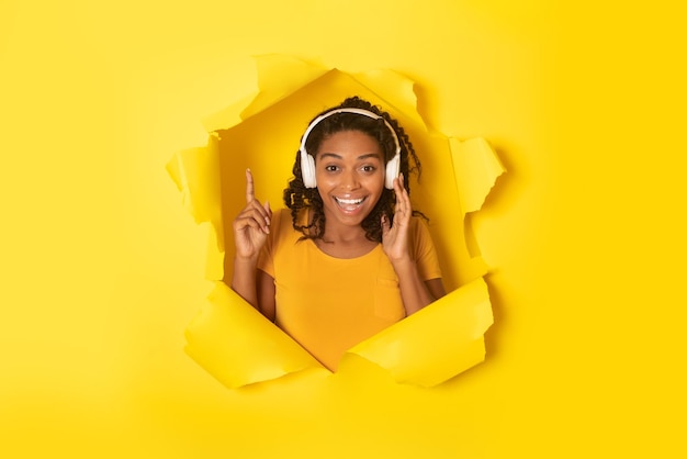 Szczęśliwa czarna dama w słuchawkach bezprzewodowych słuchająca muzyki online i patrząca na kamerę przez otwór w podartym papierze