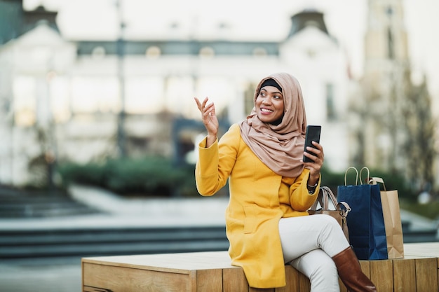 Szczęśliwa Czarna Muzułmanka Nosząca Hidżab I Prowadząca Wideorozmowę Za Pomocą Smartfona, Siedząc W środowisku Miejskim.