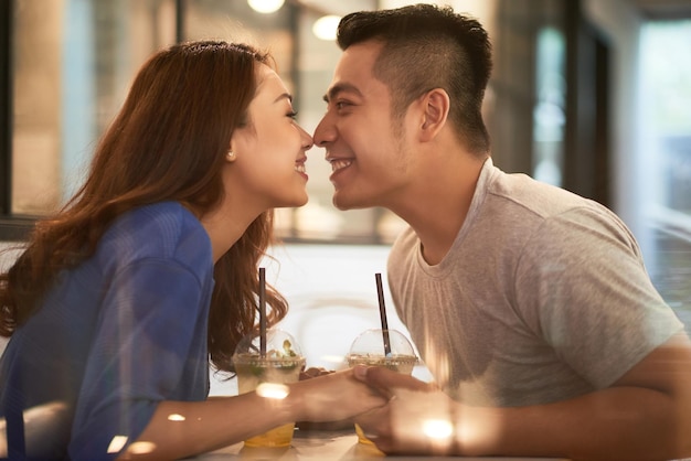 Szczęśliwa czuła młoda Azjatycka para siedzi z letnimi koktajlami i dotyka nosami nad stołem w kawiarni