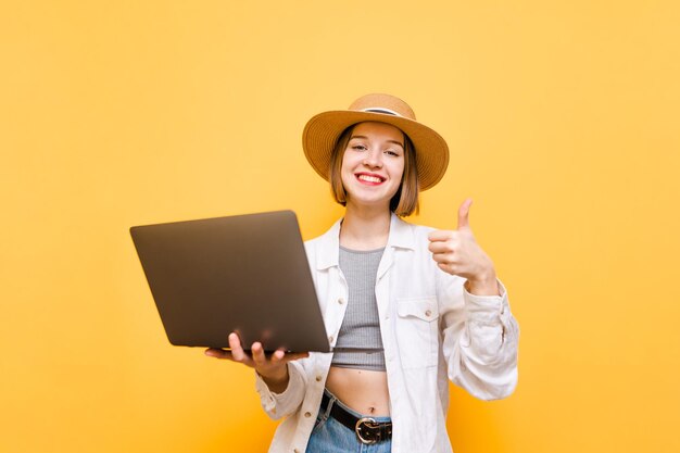 szczęśliwa dama i lekkie letnie ubrania i kapelusze stoją na żółtym tle z laptopem w rękach