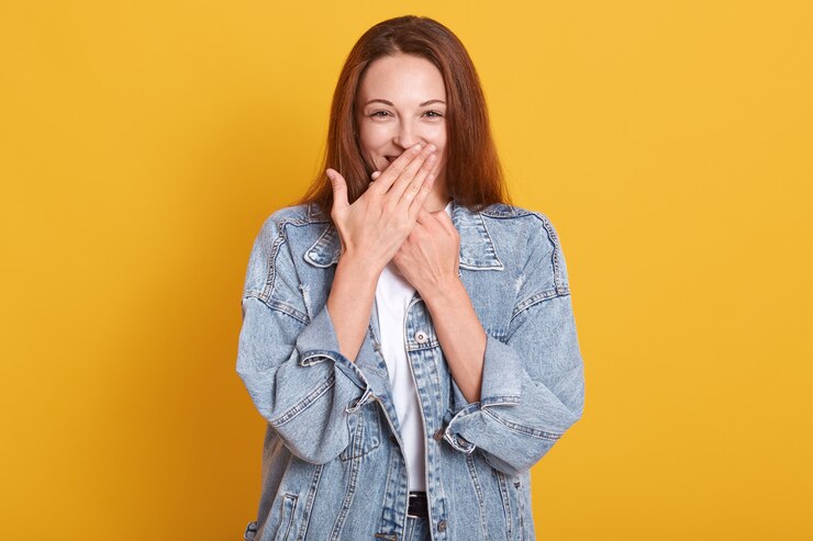 Zdjęcie szczęśliwa, dobrze wyglądająca młoda modelka zakrywająca usta obiema rękami, nieśmiała, zaskoczona, będąca w świetnym humorze, ubrana w dżinsową kurtkę i białą koszulkę, na białym tle nad żółtym