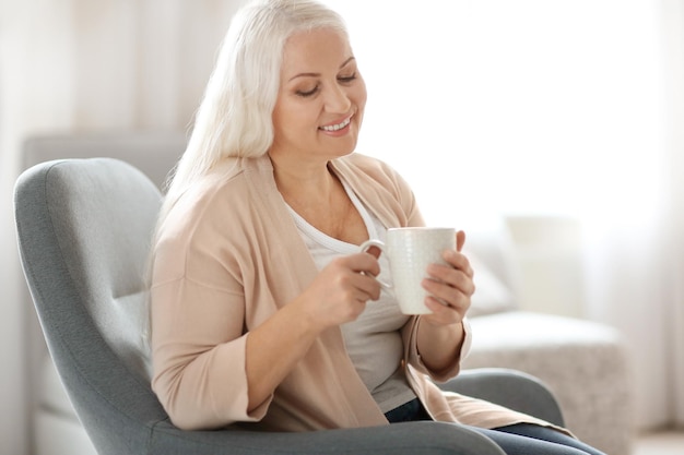 Szczęśliwa dojrzała kobieta pije herbatę siedząc w fotelu w domu