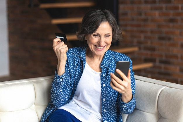 Szczęśliwa dojrzała nowoczesna kobieta zakupy online za pomocą telefonu komórkowego i karty kredytowej, siedząc na kanapie w domu. Koncepcja technologii
