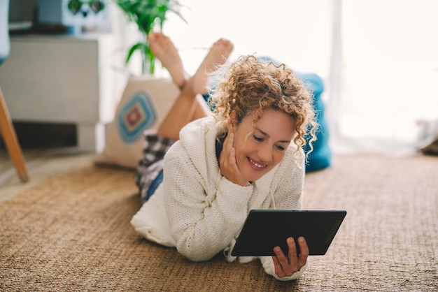 Szczęśliwa dorosła kobieta leżąc na podłodze dywan w domu przy użyciu nowoczesnego tabletu i bezprzewodowego połączenia z Internetem w domu. Kobiety cieszą się relaksem w pomieszczeniach, samotnie oglądając urządzenie internetowe