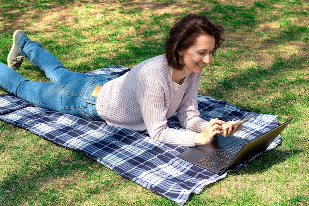 Zdjęcie szczęśliwa dorosła kobieta leży na trawie w pobliżu laptopa trzyma telefon komórkowy atrakcyjna uśmiechnięta kobieta odpoczywa