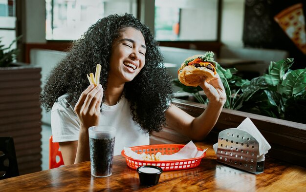 Szczęśliwa dziewczyna delektująca się hamburgerem w restauracji Afro młoda kobieta delektująca się hamburgerem z frytkami w restauracji