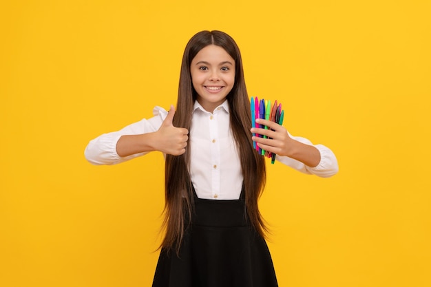 Szczęśliwa dziewczyna dziecko trzyma pisaki pokazujące kciuk w górę gest ręki na żółtym tle zadowolony