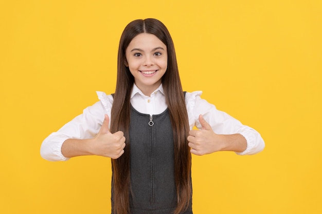 Szczęśliwa dziewczyna dziecko w szkolnym mundurku uśmiech pokazuje podwójne kciuki w górę gest dłoni na żółtym tle zadowolenie