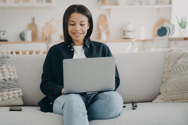 Szczęśliwa dziewczyna pisze na komputerze i uśmiecha się Freelancer siedzi na wygodnej kanapie z laptopem