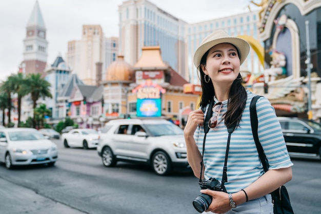 Szczęśliwa dziewczyna podróżuje w letnim pasie Las Vegas. uśmiechnięta młoda kobieta backpacker trzymając aparat vintage, patrząc na bok. kobiece stoisko turystyczne obok ruchliwej drogi z samochodami i wieżowcami na tle.