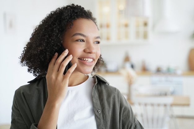 Szczęśliwa dziewczyna rasy mieszanej odpowiada na połączenie, rozmawiając przez telefon, ciesz się przyjemną rozmową mobilną w domu
