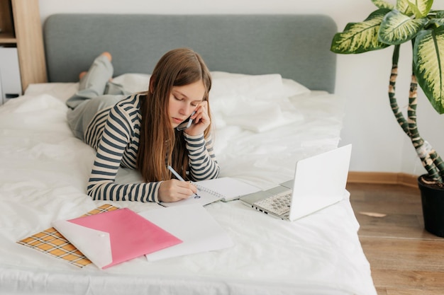 Szczęśliwa dziewczyna rozmawia przez telefon leżąc w łóżku i używając laptopa online edukacja zadania domowe