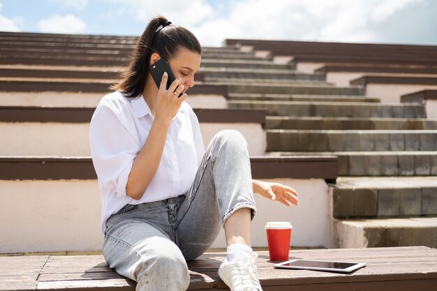 Szczęśliwa dziewczyna rozmawia przez telefon trzymając kubek papierowy kawy ciesząc się słoneczny dzień siedząc w amfiteatrze.