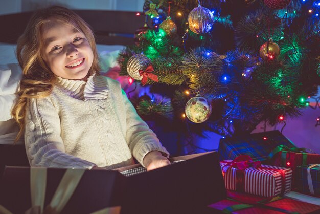 Szczęśliwa dziewczyna rozpakowuje prezent na tle choinki. wieczorna pora nocna