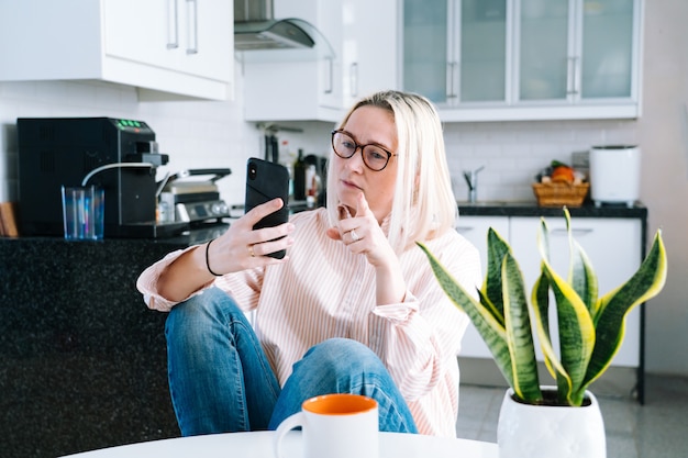 Szczęśliwa dziewczyna siedzi w domu kuchnię i trzyma połączenie wideo. Młoda kobieta używa smartphone dla rozmowy wideo z przyjacielem lub rodziną. Webinarium z nagraniem Vloggera. Kobiety przyglądająca kamera i machać powitanie ręki