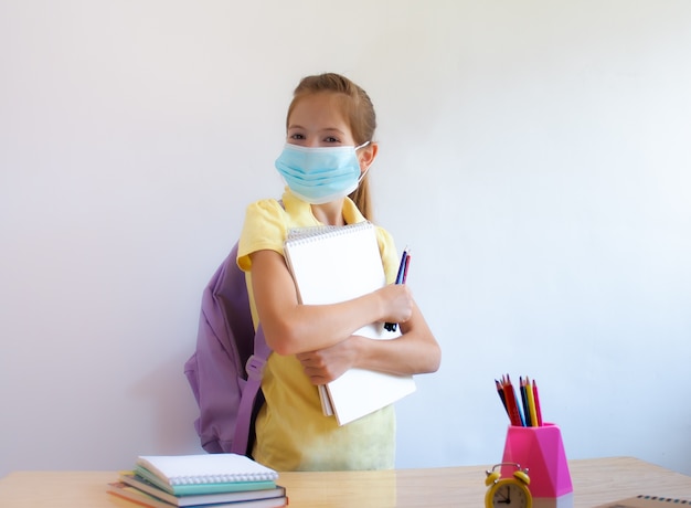 Szczęśliwa dziewczyna ubrana w ochronną maskę medyczną na twarzy trzyma przybory szkolne w dłoniach