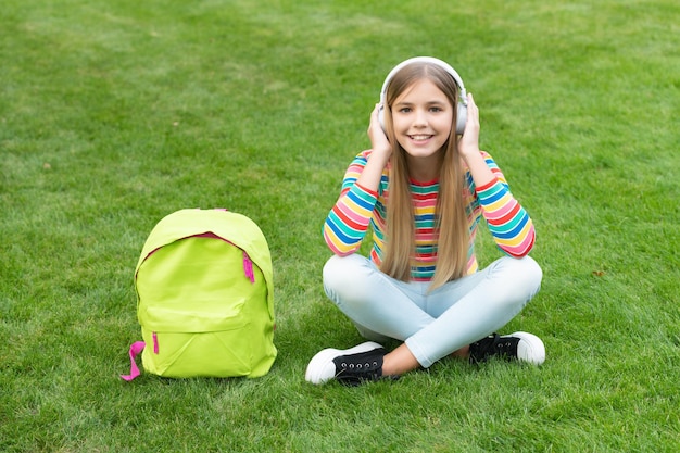 Szczęśliwa dziewczyna ucząca się zdalnie słuchając kursu audio w słuchawkach siedząc na trawie w szkole