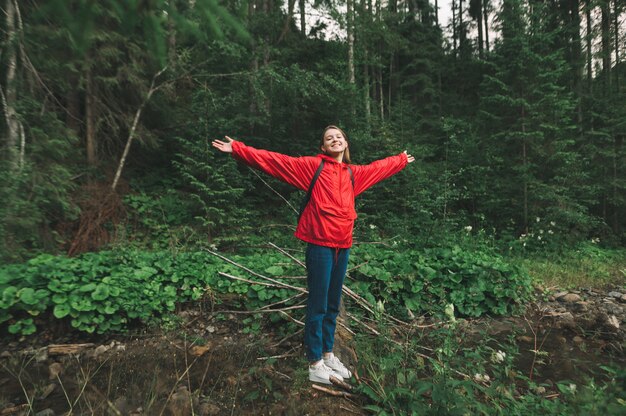 Szczęśliwa dziewczyna w czerwonym płaszczu stoi na dzienniku nad górskim potokiem