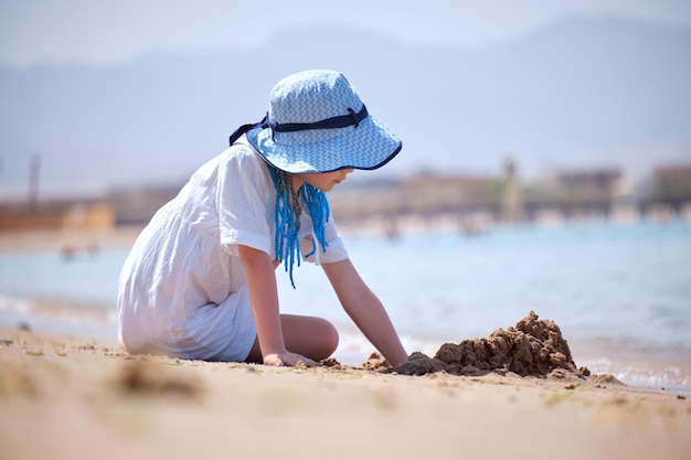 Szczęśliwa dziewczyna w dużym kapeluszu i białej sukience bawiąca się sama z mokrym piaskiem na piaszczystej plaży w pobliżu czystej wody lagun