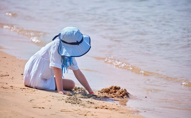 Szczęśliwa dziewczyna w dużym kapeluszu i białej sukience bawiąca się sama z mokrym piaskiem na piaszczystej plaży w pobliżu czystej wody lagun