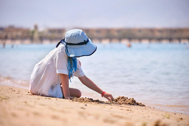 Szczęśliwa dziewczyna w dużym kapeluszu i białej sukience bawiąca się sama z mokrym piaskiem na piaszczystej plaży w pobliżu czystej wody lagun