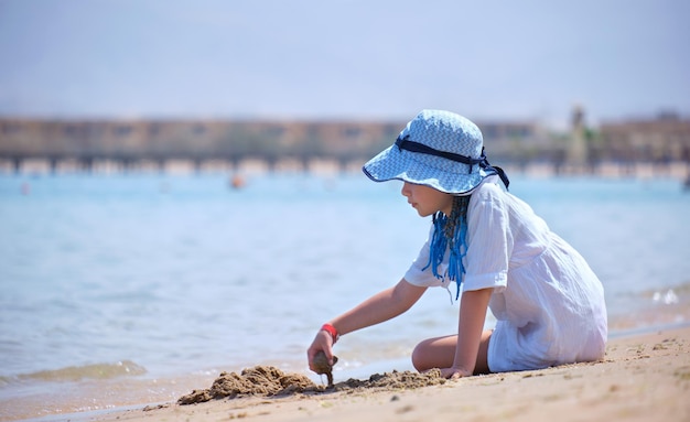 Szczęśliwa dziewczyna w wielkim kapeluszu i białej sukni bawi się samotnie z mokrym piaskiem na piaszczystej plaży w pobliżu czystej wody w lagunie morskiej