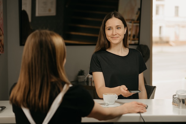 Szczęśliwa dziewczyna z długimi włosami uśmiecha się i płaci za kawę smartfonem dzięki bezdotykowej technologii PAY PASS w kawiarni. Brunetka-barista trzyma terminal za płacenie klientowi.