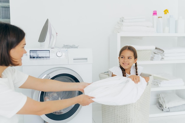 Szczęśliwa dziewczyna z dwoma warkoczykami pozuje w koszu z brudną pościelą bawi się w pralni z mamą pomaga w praniu Kobieta ładuje pralkę spędza weekend w domu zajęta pracami domowymi