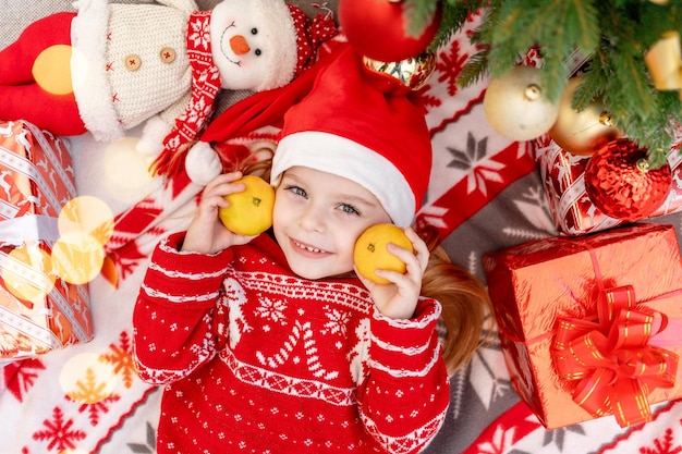 Szczęśliwa dziewczynka leży w domu pod choinką z mandarynkami i czeka na nowy rok lub Boże Narodzenie w czerwonym kapeluszu Świętego Mikołaja, uśmiecha się i raduje