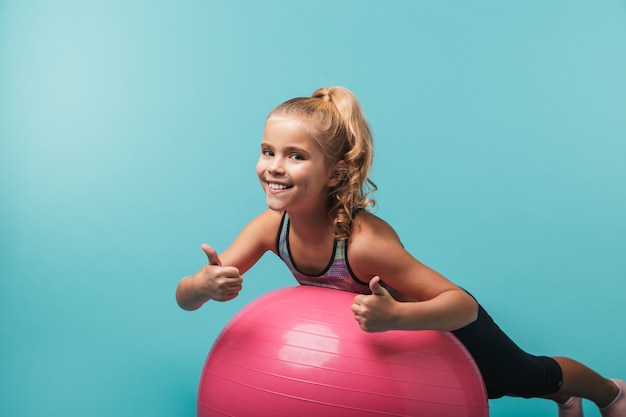 Szczęśliwa dziewczynka na sobie odzież sportową ćwiczeń z piłką fitness na białym tle nad niebieską ścianą