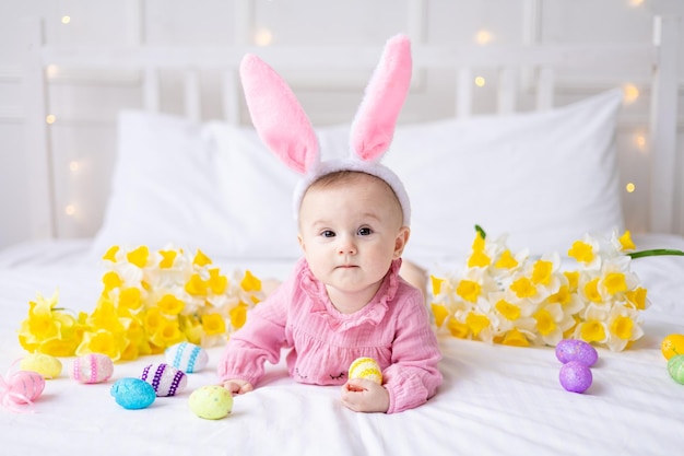 Szczęśliwa dziewczynka rasy kaukaskiej z uszami królika na głowie leży na łóżku w domu z kolorowymi jajkami wielkanocnymi i żółtymi wiosennymi kwiatami patrzy w kamerę śmiejąc się Wielkanocne dziecko