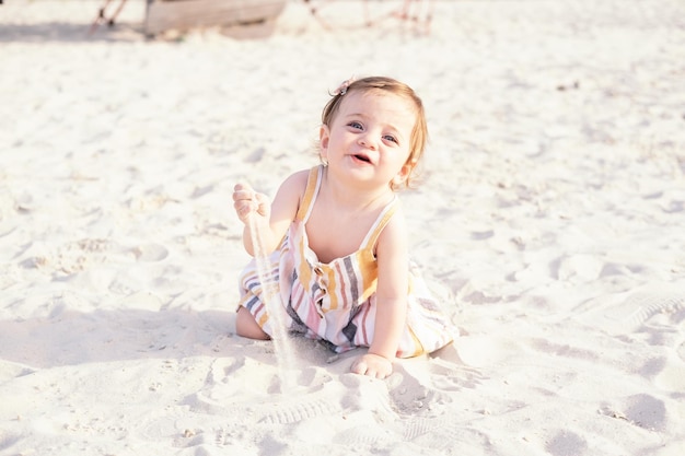 szczęśliwa dziewczynka siedzi na plaży na piasku ubrana w letnią sukienkę w paski w słoneczny dzień